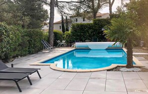 Ein rechteckiger Pool mit einer gefliesten Terrasse, umgeben von Bäumen und einem weißen Gebäude im Hintergrund.