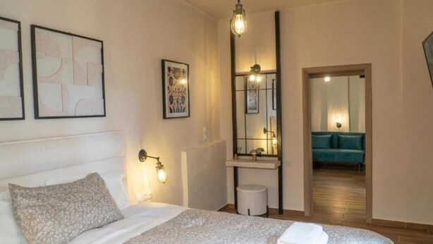 Ein Schlafzimmer mit Bett, Nachttisch, Wandlampe, Spiegel und Blick in einen angrenzenden Raum mit Sofa.