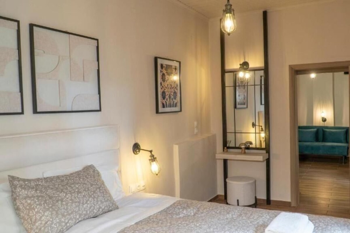 Un dormitorio con cama, mesita de noche, luz de pared, espejo y vista a otra habitación con sofá.