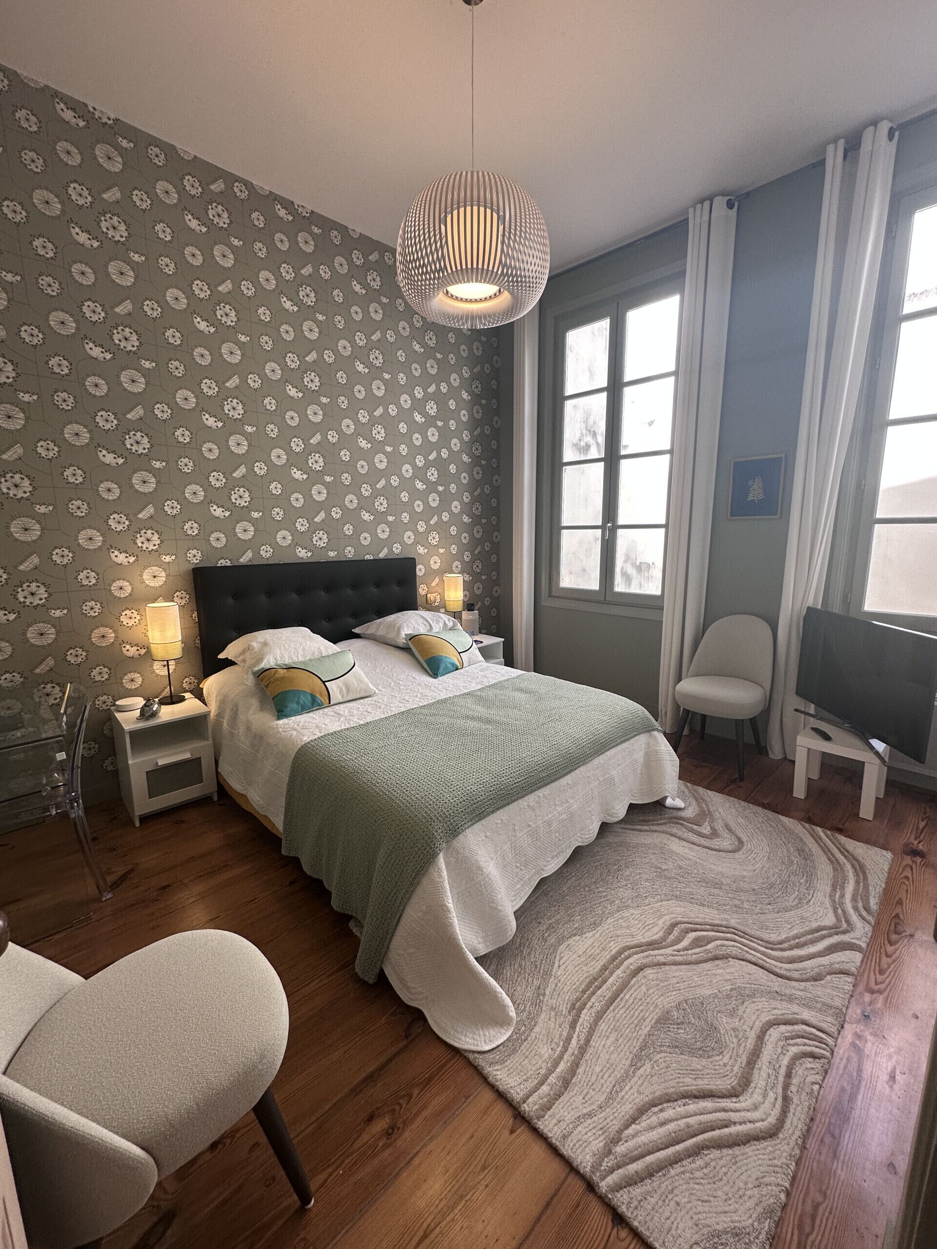 Un dormitorio con cama, lámparas de noche, televisor, una silla y un papel tapiz estampado.