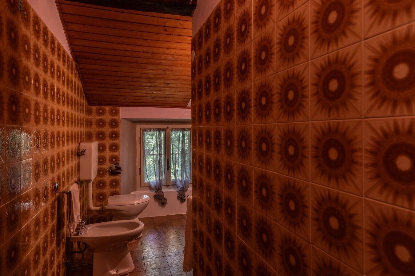 Ein Badezimmer mit gemusterten Wänden, Holzboden und zwei Toiletten, sowie ein Fenster mit Vorhängen.