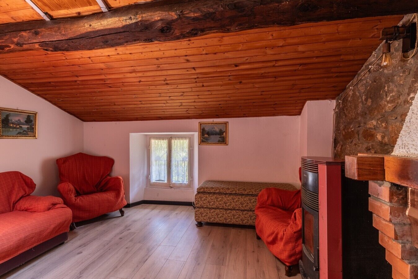 Una habitación con vigas de madera en el techo, dos sillones rojos, un sofá estampado, una chimenea y una ventana con cortinas.
