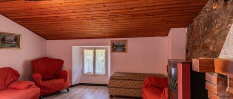 Una habitación con vigas de madera en el techo, dos sillones rojos, un sofá estampado, una chimenea y una ventana con cortinas.