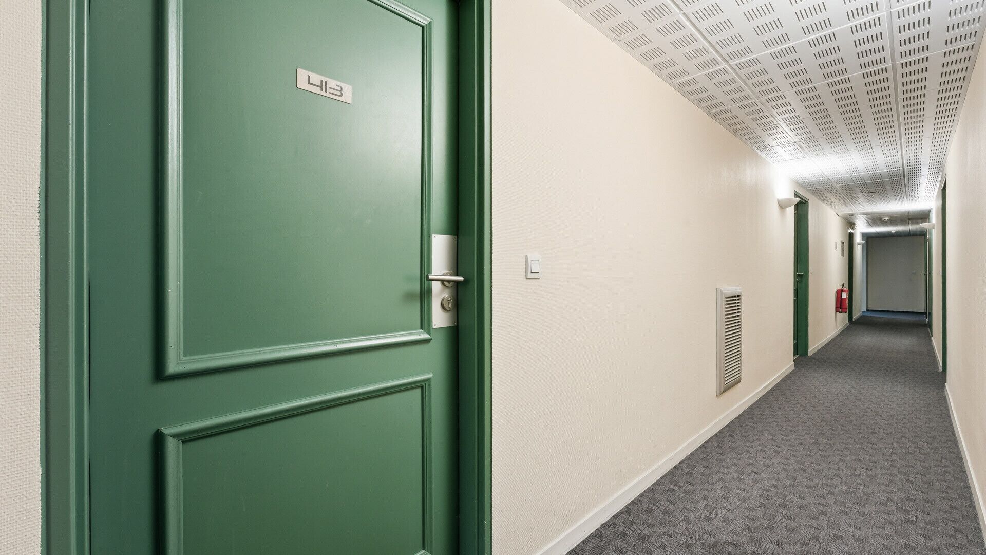 Un pasillo con una puerta verde, un interruptor de luz, una rejilla de ventilación y un extintor.