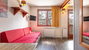 Une pièce de taille modeste, avec un canapé rouge, une petite table, un téléviseur fixé au mur, une fenêtre avec des rideaux et une vue sur les bâtiments environnants.