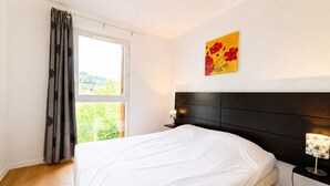 Une chambre à coucher avec un lit, une table de chevet, une lampe, une fenêtre avec des rideaux et un tableau au mur.