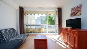 Quarto com sofá, televisão sobre um armário de madeira, uma mesinha e varanda com uma cadeira.