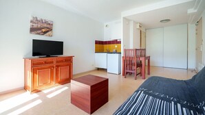 Uma sala de estar com televisão, sofá, armário de madeira, mesa pequena e área de cozinha com frigorífico e uma cadeira.