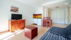 A living room with a television, a sofa, a wooden cabinet, a small table, and a kitchen area with a refrigerator and a chair. - Charming Studio - Sleeps 2 - Close to the sea (Agde)