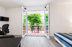 Ein Zimmer mit einem Bett, einem Schreibtisch, einem Sessel und einem Tisch mit Stühlen. An der Wand ist ein Fernseher angebracht und eine Glastür führt zu einem Außenbereich mit Tisch und Stühlen.
