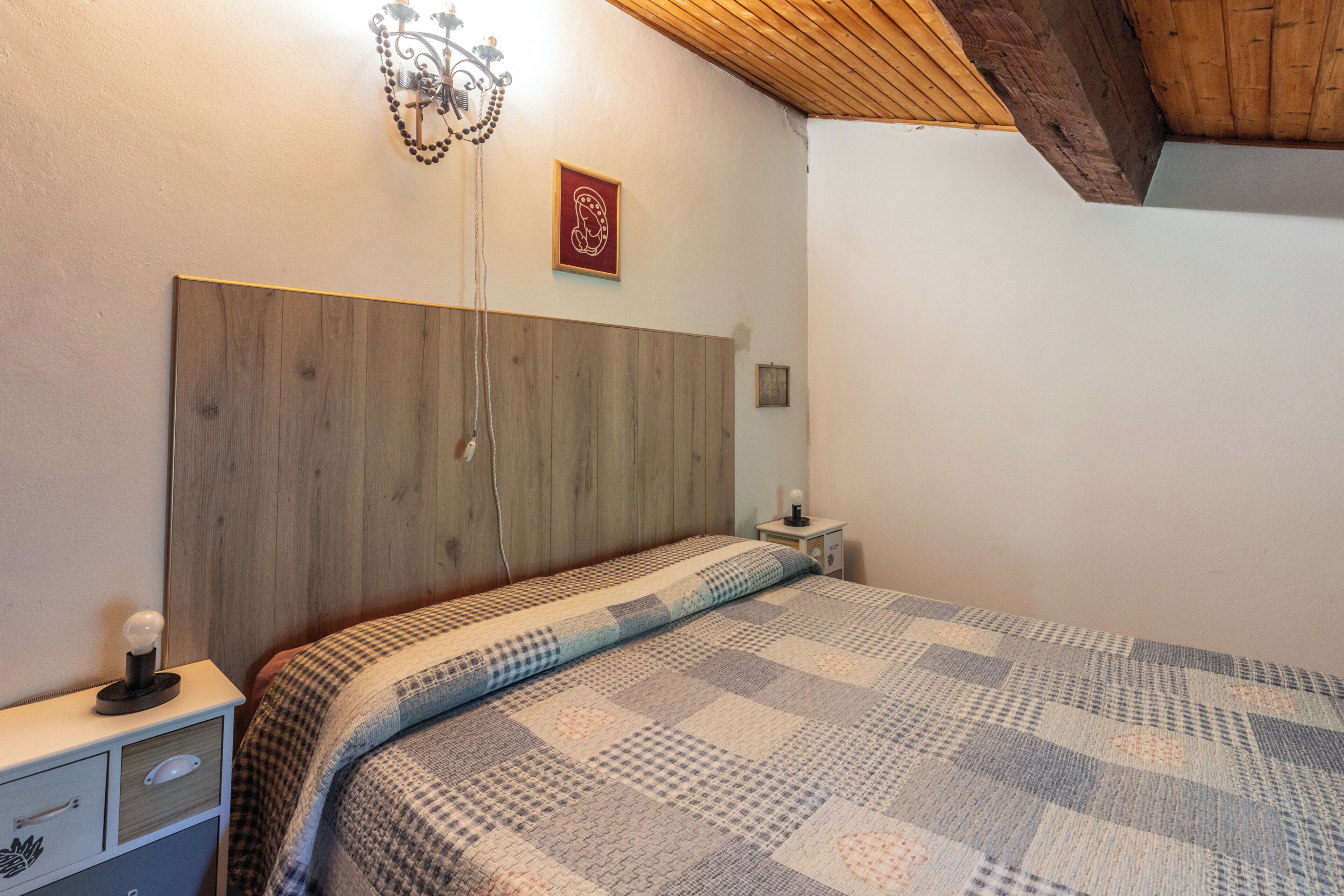 A bedroom with a wooden headboard, a patterned bedspread, a bedside table with a lamp, and a wall-mounted light fixture.
