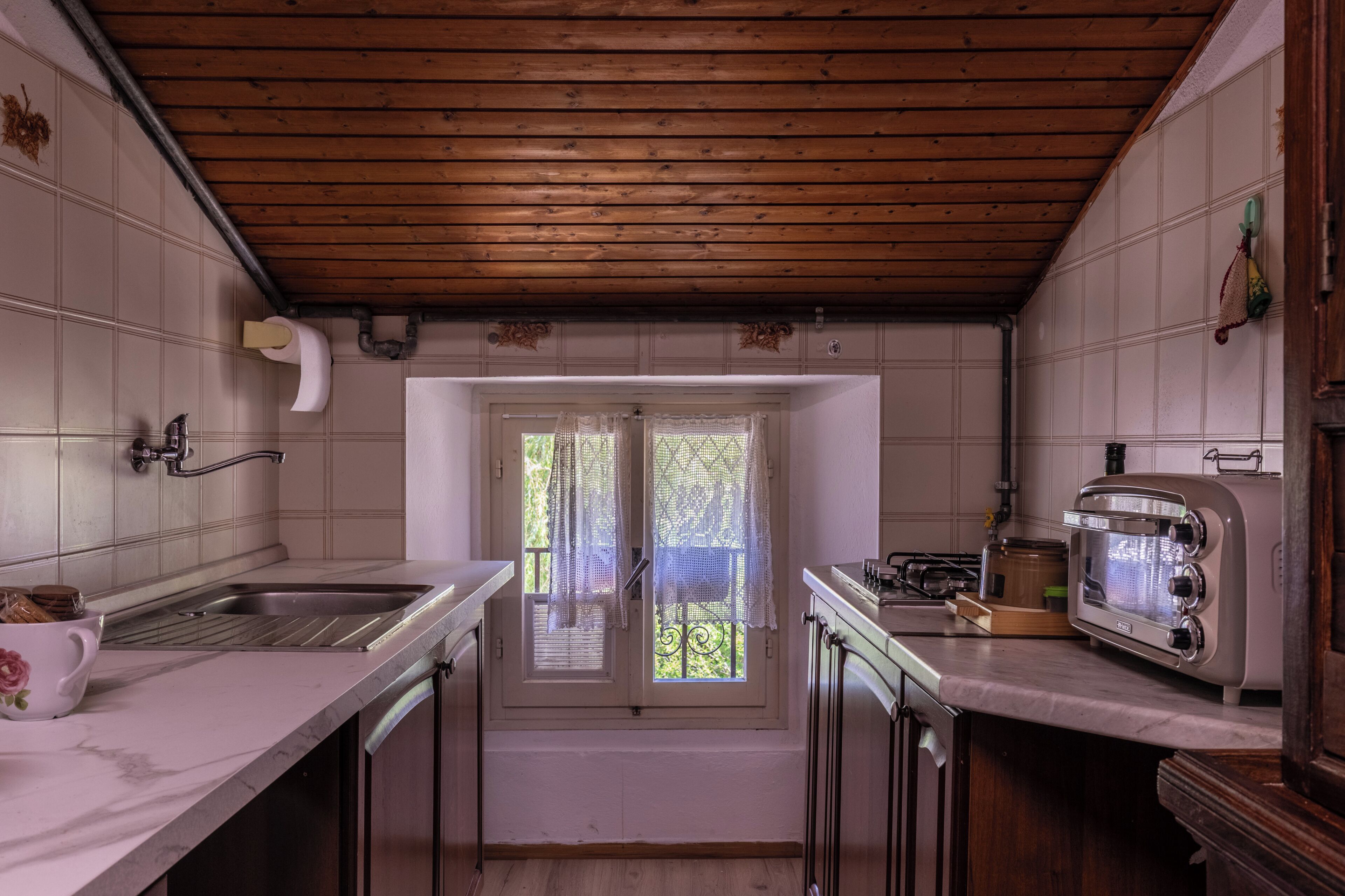 Eine Küche mit Holzbalkendecke, einem Fenster mit Spitzenvorhängen, einer Spüle, einem Herd und einem alten Toaster.