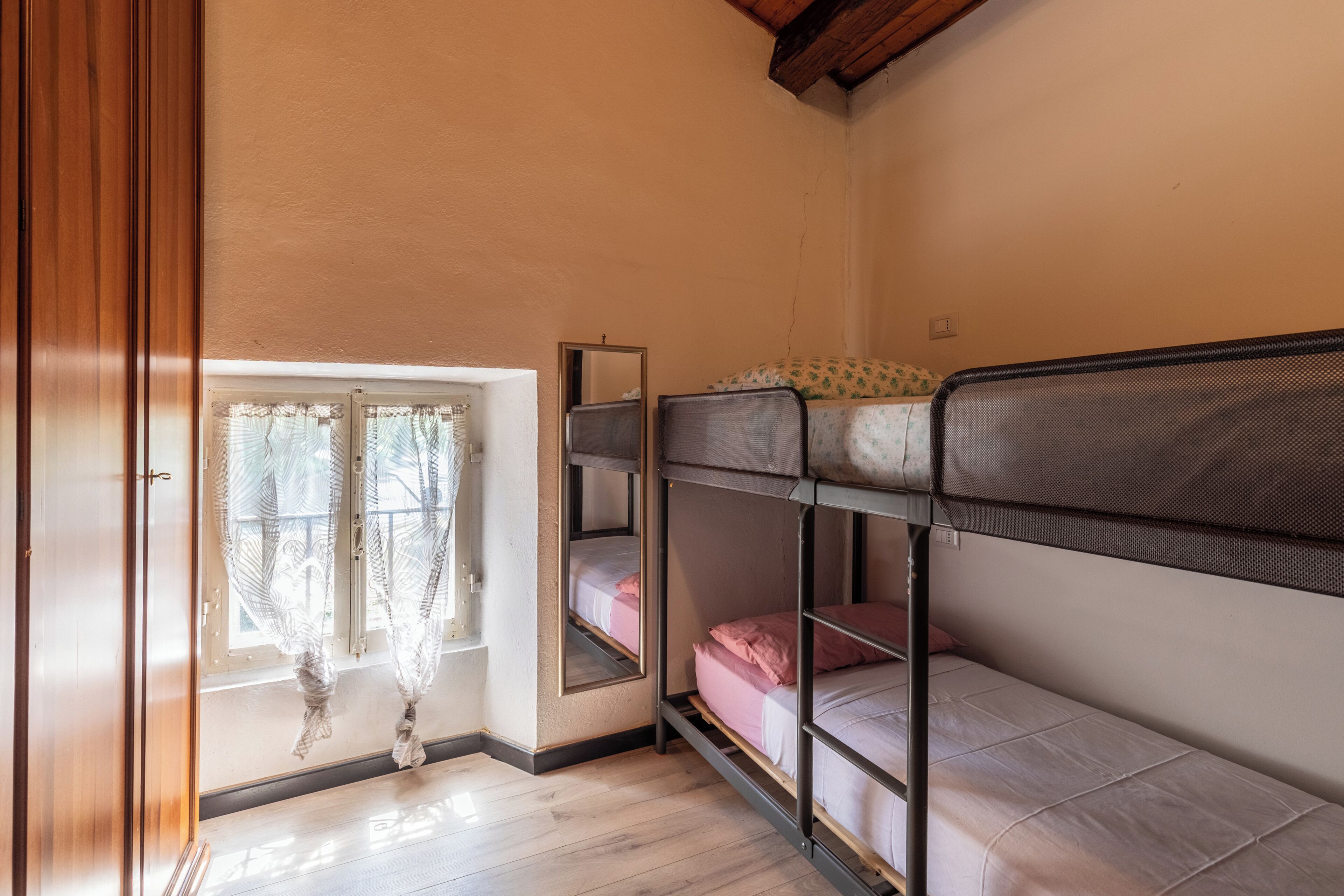 A dormitory room with bunk beds, a window with curtains, and a wooden floor.