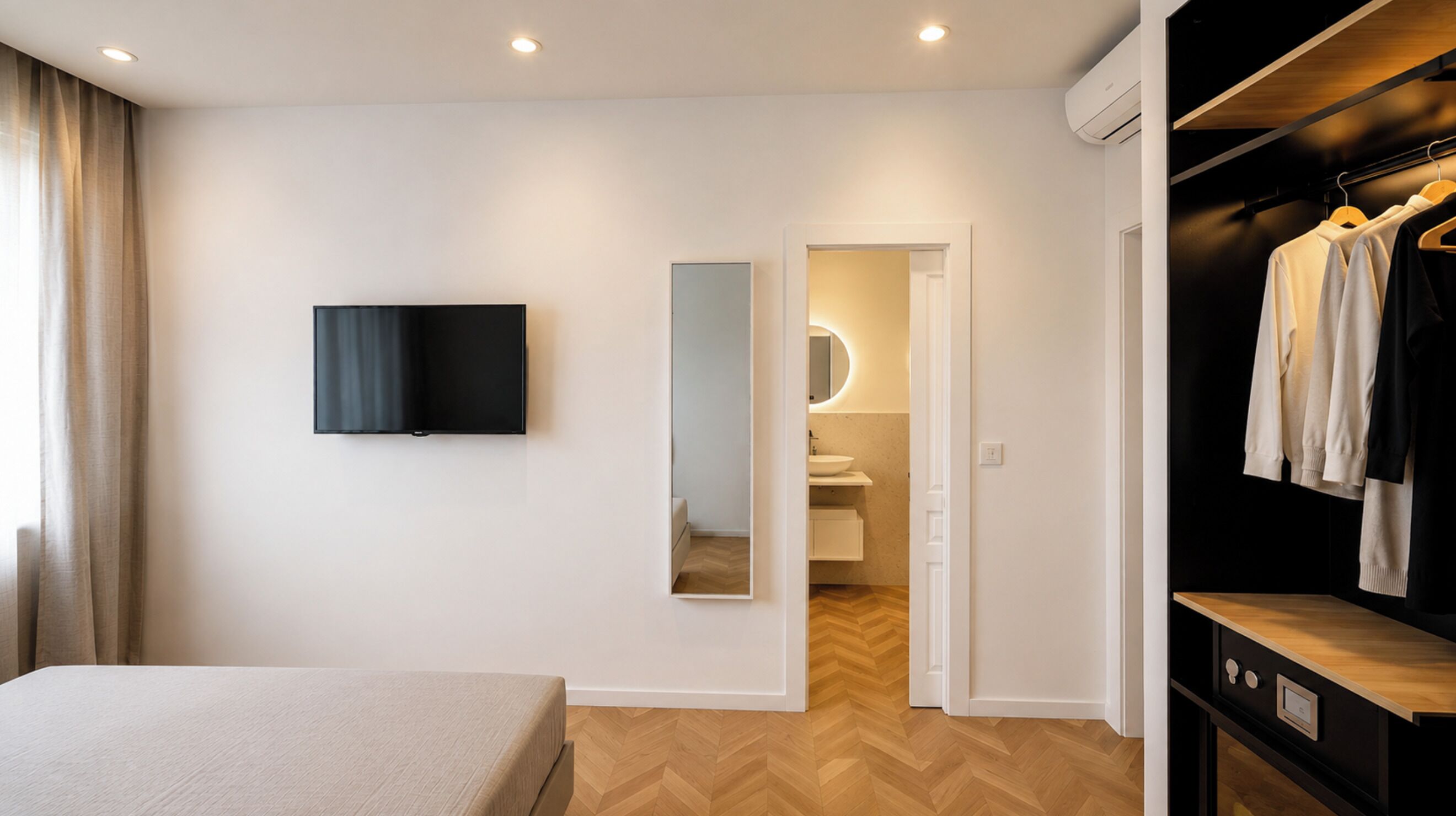 A modern hotel room with a bed, a TV, a mirror, a wardrobe, and a bathroom visible through an open door.