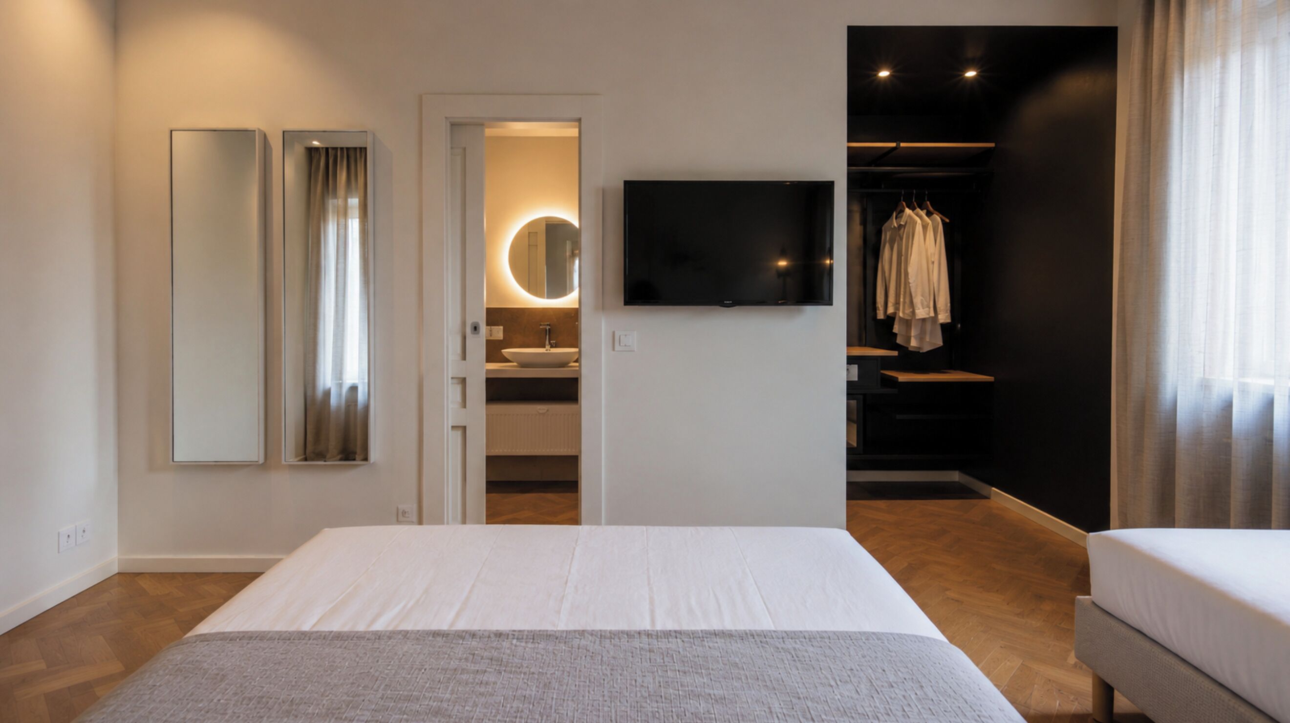 A hotel room with a bed, a TV, a mirror, a bathroom with a sink, and a closet with clothes.
