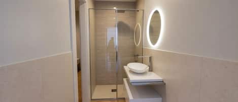 Un baño moderno con ducha envuelta en vidrio, un lavamanos blanco y dos espejos ovalados con luz.