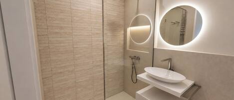 Un baño moderno con ducha envuelta en vidrio, un lavabo blanco y dos espejos adosados a la pared con iluminación integrada.