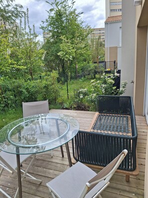 Mesa de vidro e duas cadeiras de vime em um deck de madeira, com vista para um jardim e edifícios.