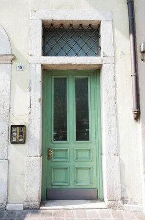 Eine grüne Tür mit einem kleinen Fenster darüber, der Hausnummer '19' an der Wand und einem Metallgitter vor dem Fenster.