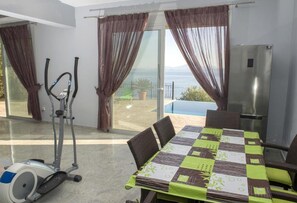 Een kamer met een eettafel, stoelen, een fitnessapparaat, een koelkast en uitzicht op zee door grote ramen.