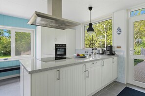 Eine moderne Küche mit weißen Schränken, einem eingebauten Ofen, einer Kaffeemaschine und einem großen Fenster mit Blick auf Grünflächen.