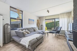 A modern living room with a sofa, a chair, a small table, and a TV. There are large windows with curtains, a fan, and a painting on the wall. - Enjoy peace, space and comfort at Haringvliet 93 in Noordzeepark Ouddorp. This charming holiday home is located close to the beach and nature, making it ideal for families, friends and dog lovers. (Ouddorp)