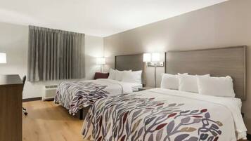 A hotel room with two beds, a desk, a chair, a window with curtains, and a wall-mounted lamp.