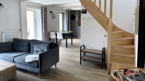 A modern living room with a grey sofa, a wooden staircase, and a dining area in the background. - Holiday Home 'Gite Baie' with Private Terrace, Private Garden and Wi-Fi (Telgruc-sur-Mer)