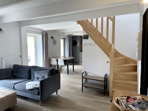 Ein modernes Wohnzimmer mit einem grauen Sofa, einer Holztreppe und einem Essbereich im Hintergrund.
