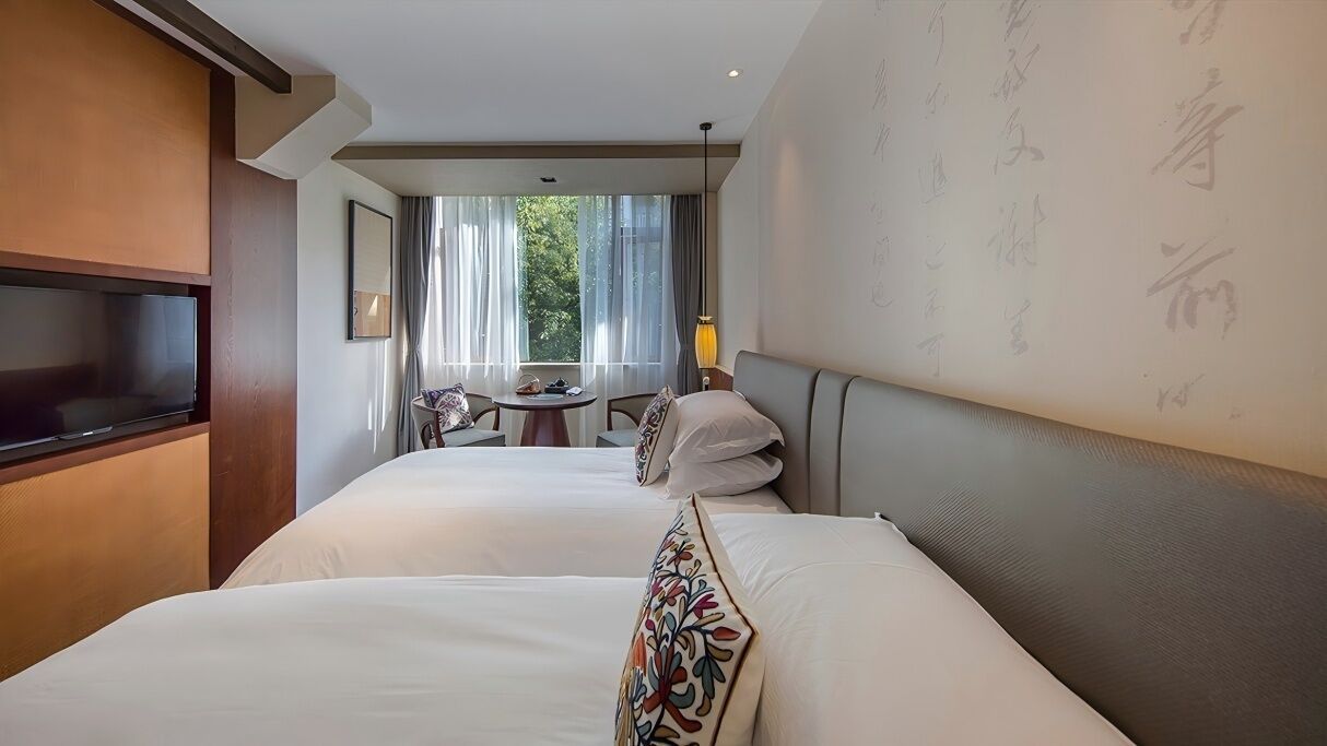 A hotel room with two beds, a TV, a small table with chairs, and a window with curtains.