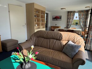 A living room with a brown leather sofa, a wooden bookshelf, a dining table with chairs, and a window with curtains. - Holiday home Bohemien in Sint Maartenszee in Callantsoog. Suitable for 6 people with 3 bedrooms. (Sint Maartensvlotbrug)