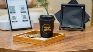 Una taza de café sobre una bandeja, con el texto 'OPEN HOTEL', un código QR enmarcado y una planta al fondo.