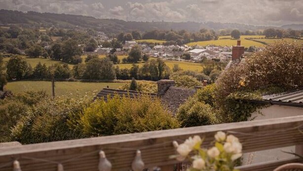 Una terraza con vistas a un paisaje rural, que incluye campos y un pueblo. Hay sillas de mimbre y una mesa con un jarrón de flores y una botella.