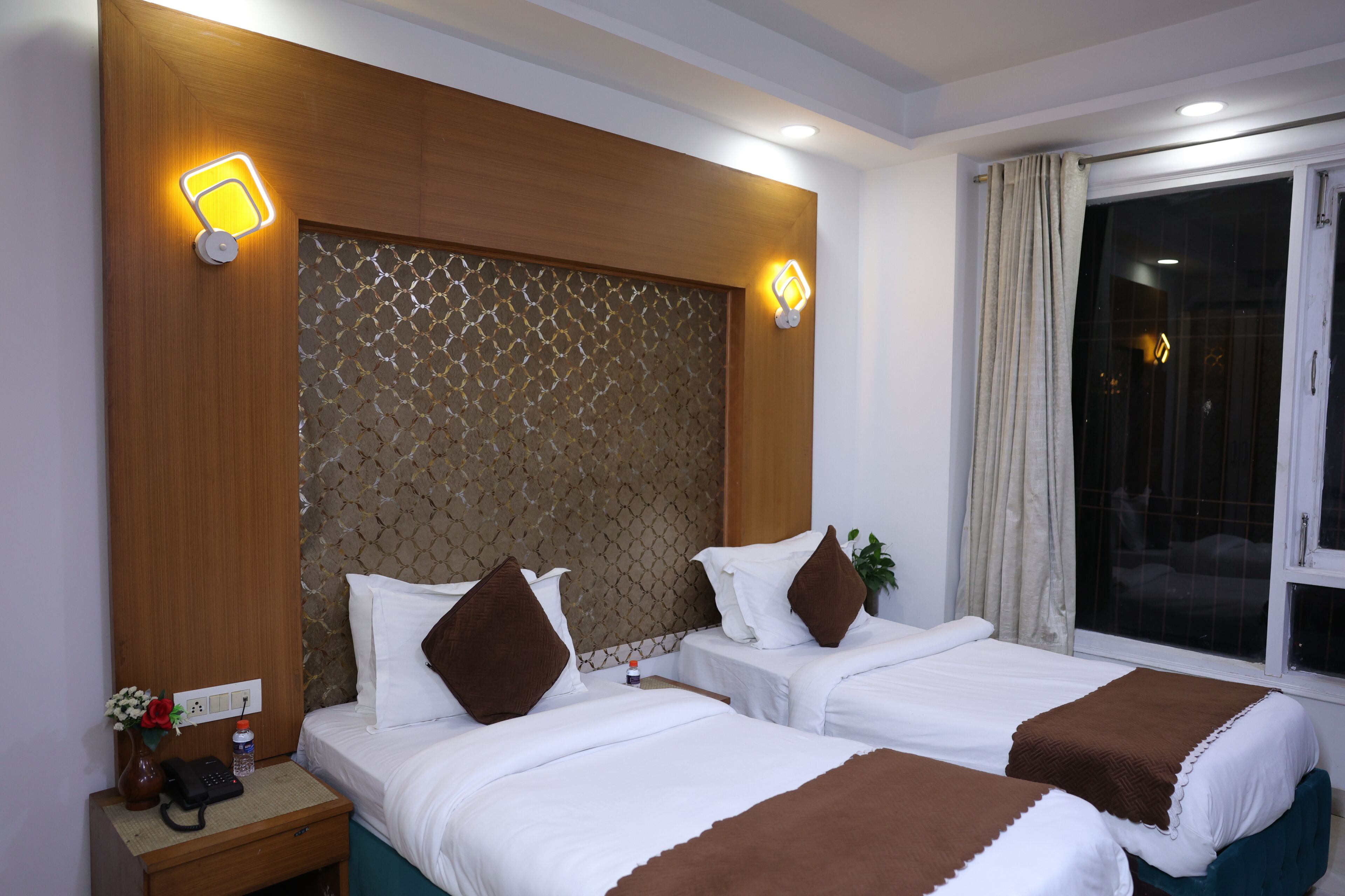 Habitación de hotel con dos camas, cabecera de madera, mesita de noche con teléfono y ventana con cortinas.