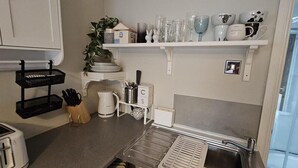 A kitchenette with a toaster, kettle, knife block, and a shelf with glassware and plates.
