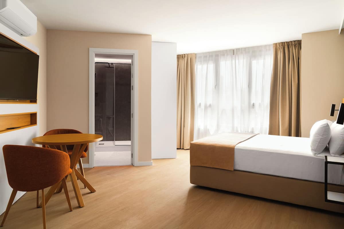 Una habitación de hotel con cama, una mesa con sillas, televisor y puerta que da a un balcón.