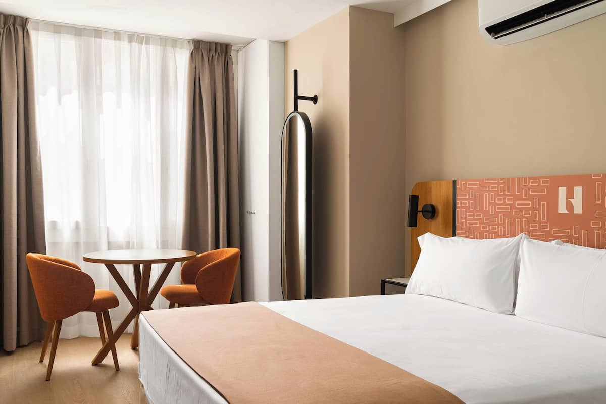 Habitación de hotel con cama, una mesa, dos sillas, mesita de noche, lámpara y equipo de aire acondicionado.
