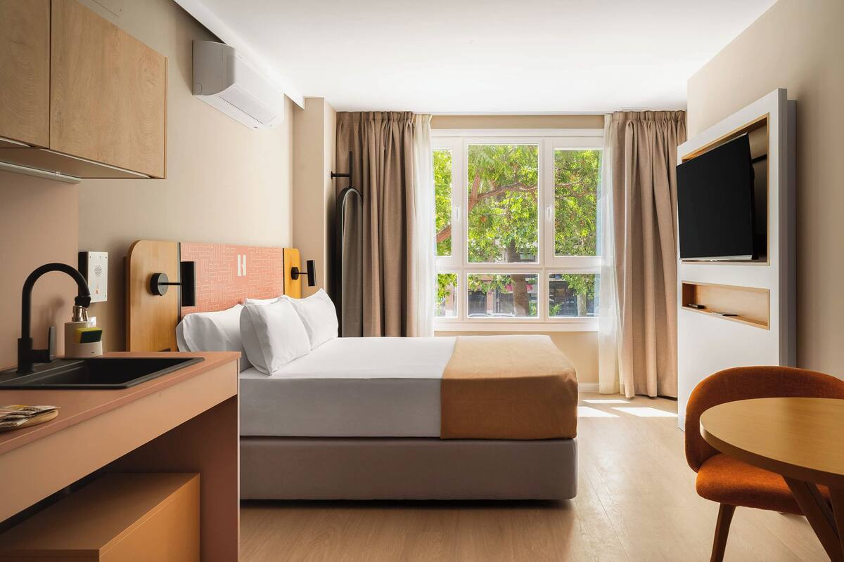 Una habitación de hotel con cama, escritorio, silla, televisor y ventana con cortinas.