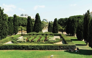 Um jardim bem cuidado, com sebes bem aparadas, árvores altas e um caminho central que leva a um edifício com estrutura em forma de cúpula.