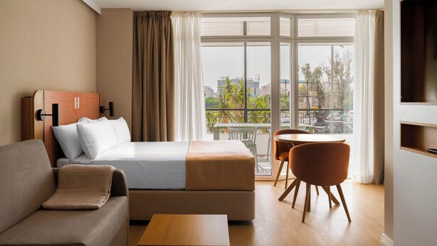 Una camera d'albergo con un letto, un divano, un tavolino, una TV e un balcone con vista sulla città.