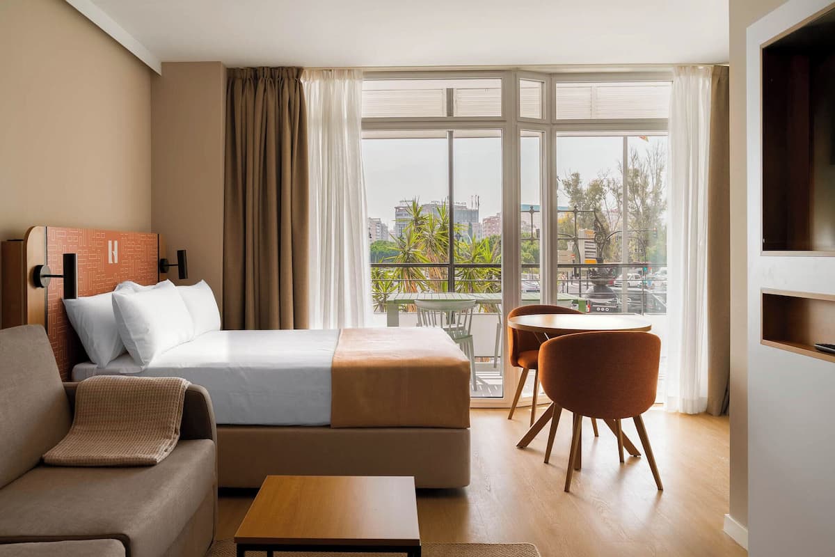 Habitación de hotel con cama, sofá, mesita, televisión y balcón con vista a la ciudad.
