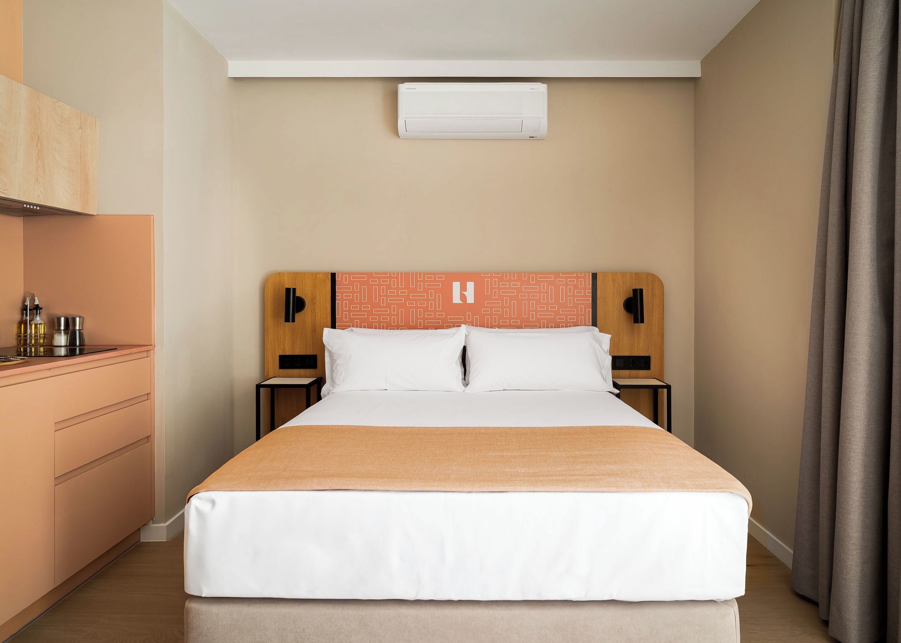 Habitación de hotel con cama, mesitas de noche, un escritorio pequeño con silla, aire acondicionado montado en la pared y una ventana con cortinas.