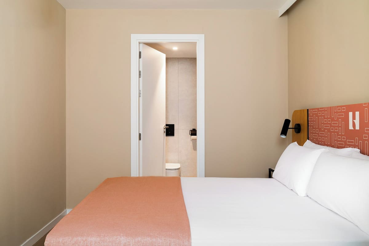 Una habitación de hotel con una cama, una puerta que da a un baño y una lámpara fijada a la pared.