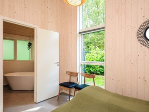 Una habitación con detalles de madera, una bañera, dos sillas y un ventanal con vistas a la vegetación.