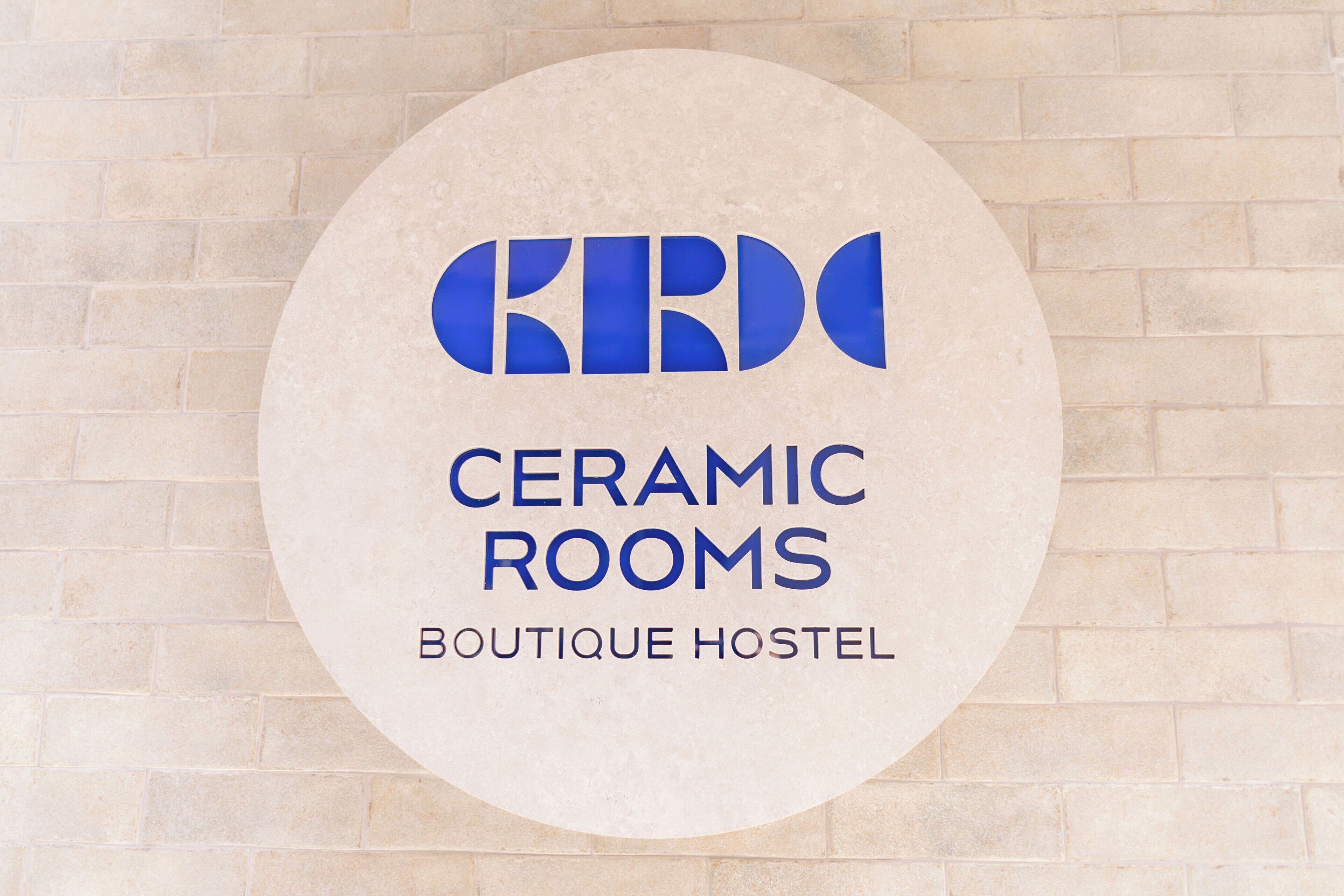 Een rond bord met de tekst 'CERAMIC ROOMS BOUTIQUE HOSTEL' voor een achtergrond van bakstenen.