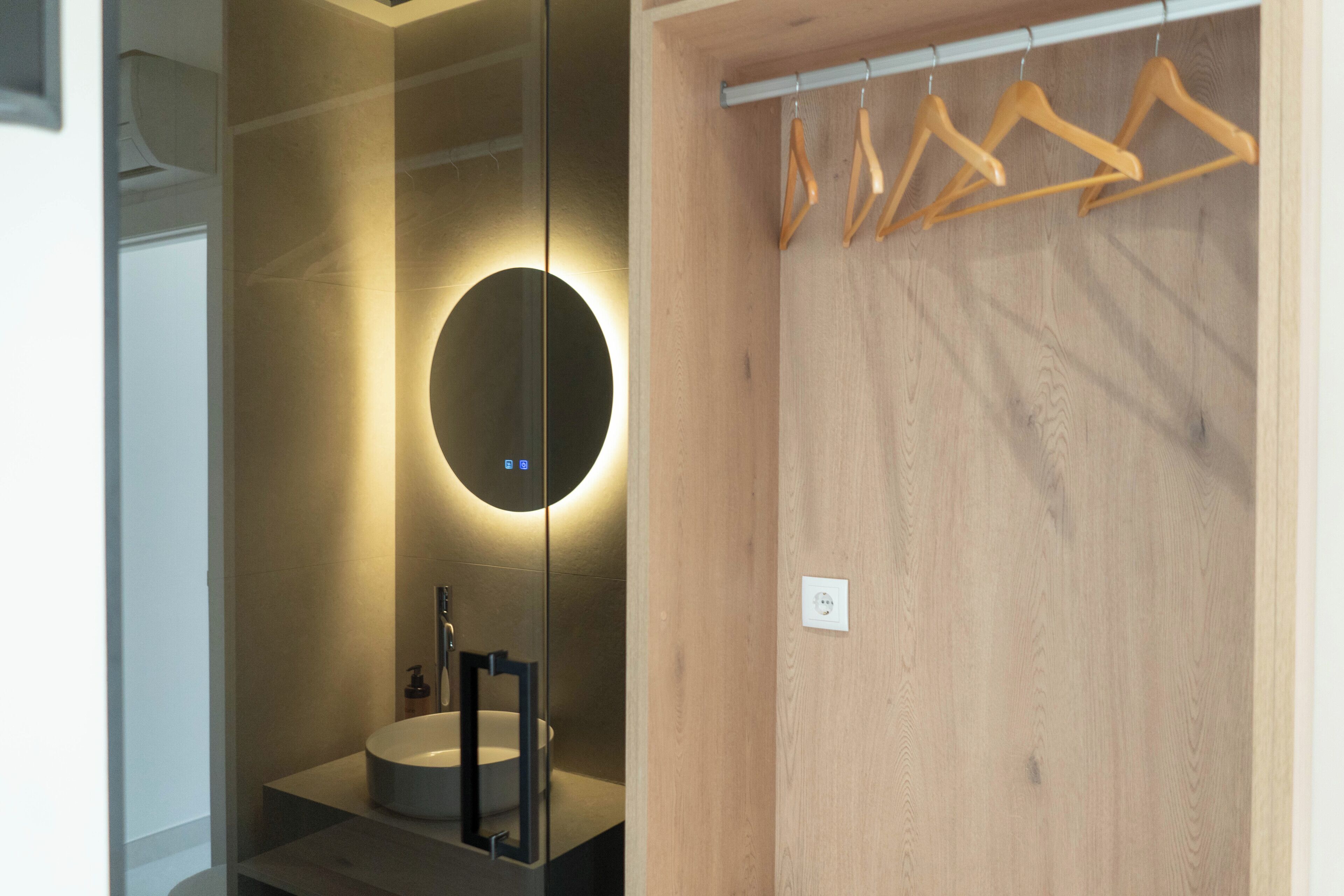 Un baño moderno con un espejo redondo, un lavamanos y perchas de madera en un perchero.