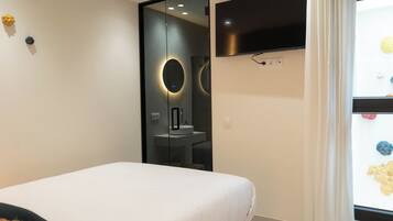 Un dormitorio moderno con cama, espejo, televisor y ventana con vistas al exterior.
