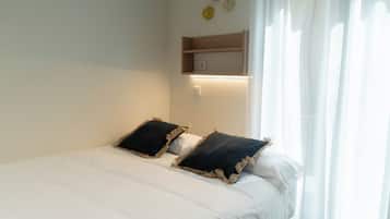 A bed with white bedding, two dark pillows with tassels, a wooden shelf above the bed, and a wall-mounted light.