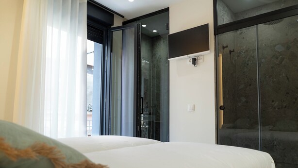 A modern hotel room with a bed, a glass shower enclosure, and a view of the outside. - Ceramic Rooms Boutique Hostel (Villarreal)