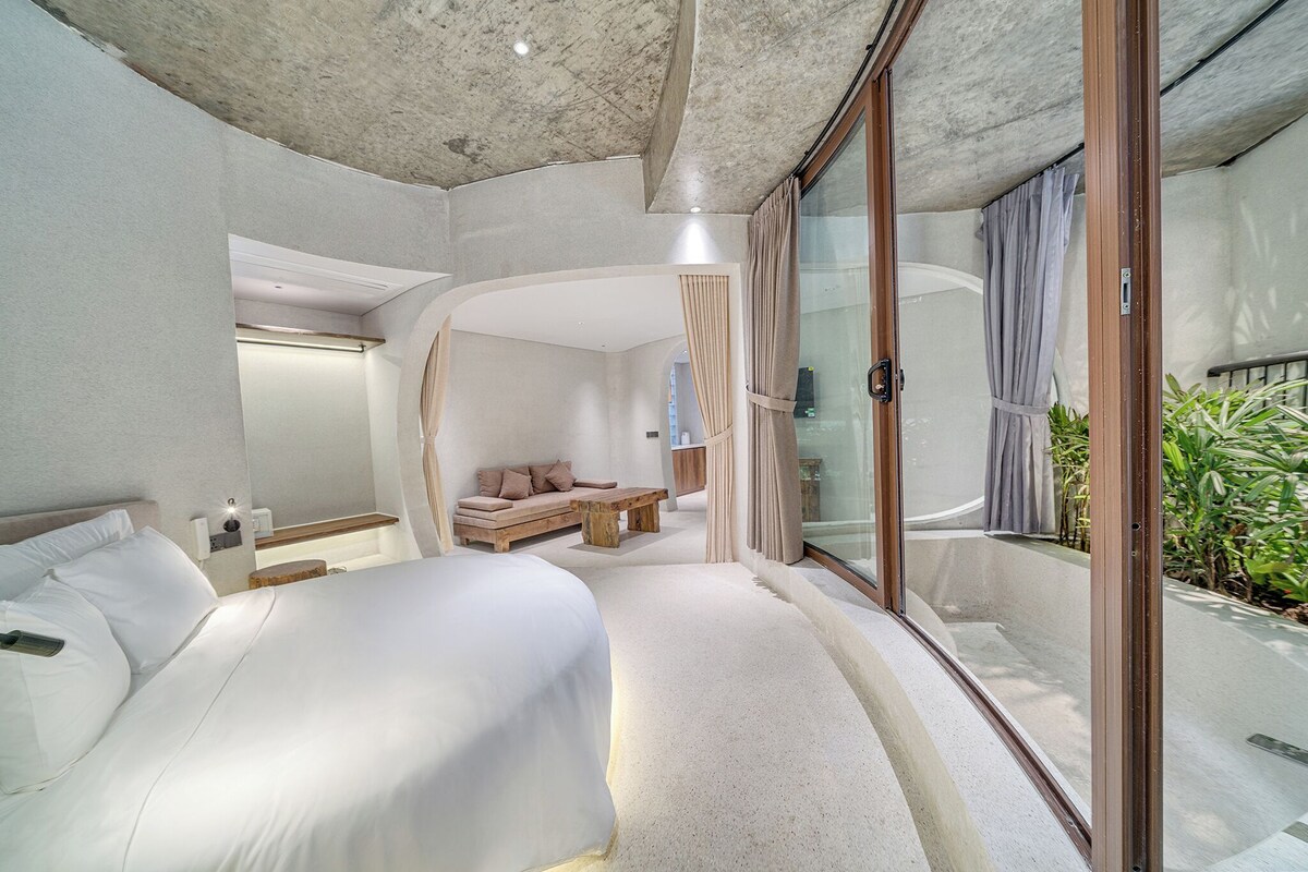 A modern hotel room with a large bed, a sofa, a small table, and a sliding glass door leading to a balcony with plants.